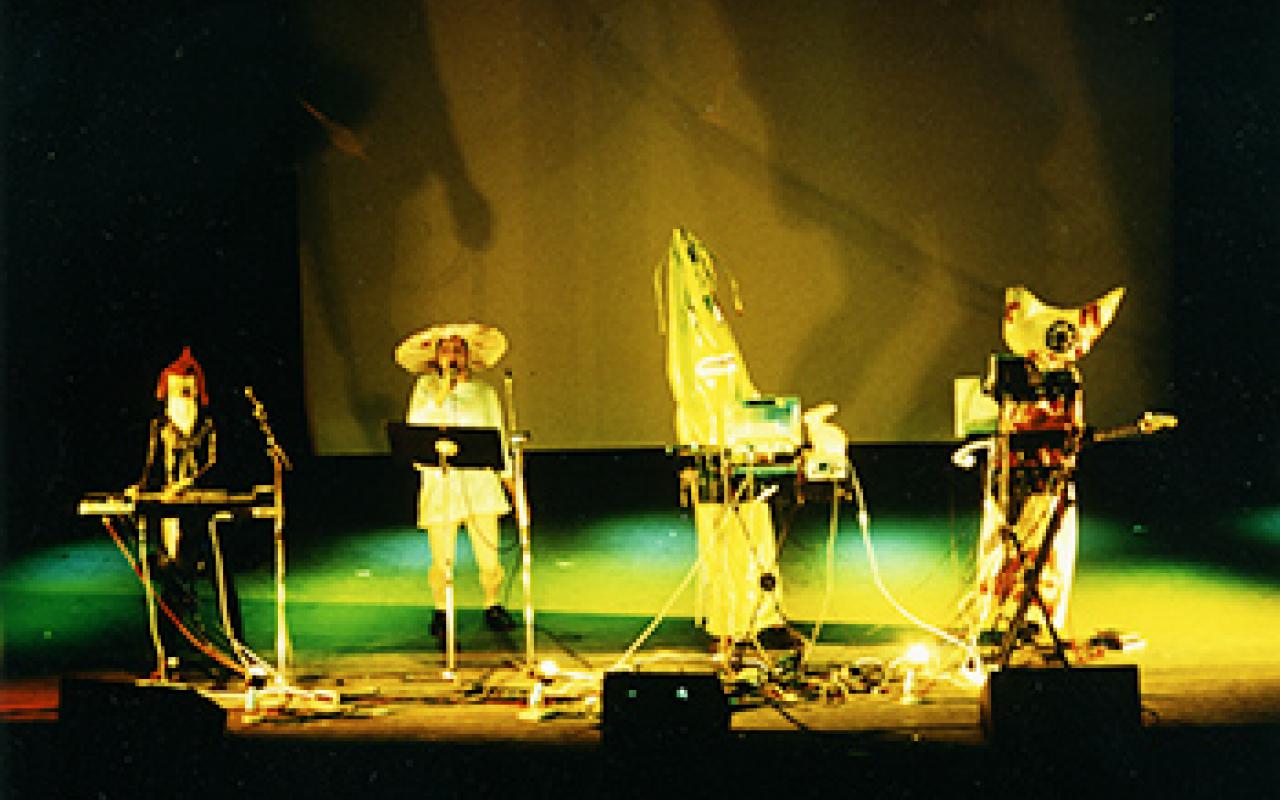 Four figures standing on a stage with colorful costumes and instruments.