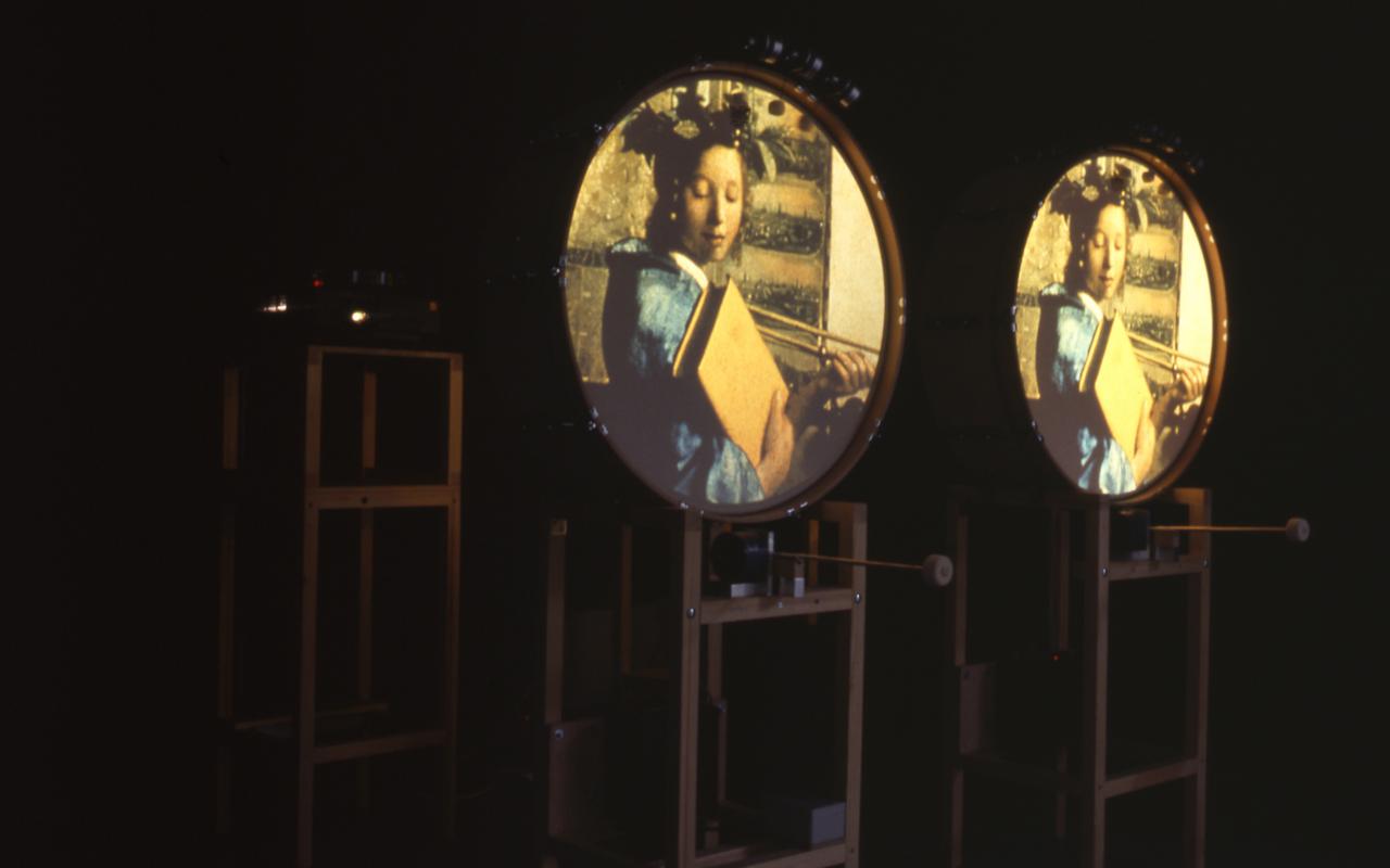 There are three racks to be seen. On two of them round surfaces are fixed. On each of them the same image is projected: a young woman with a large book in her hand.
