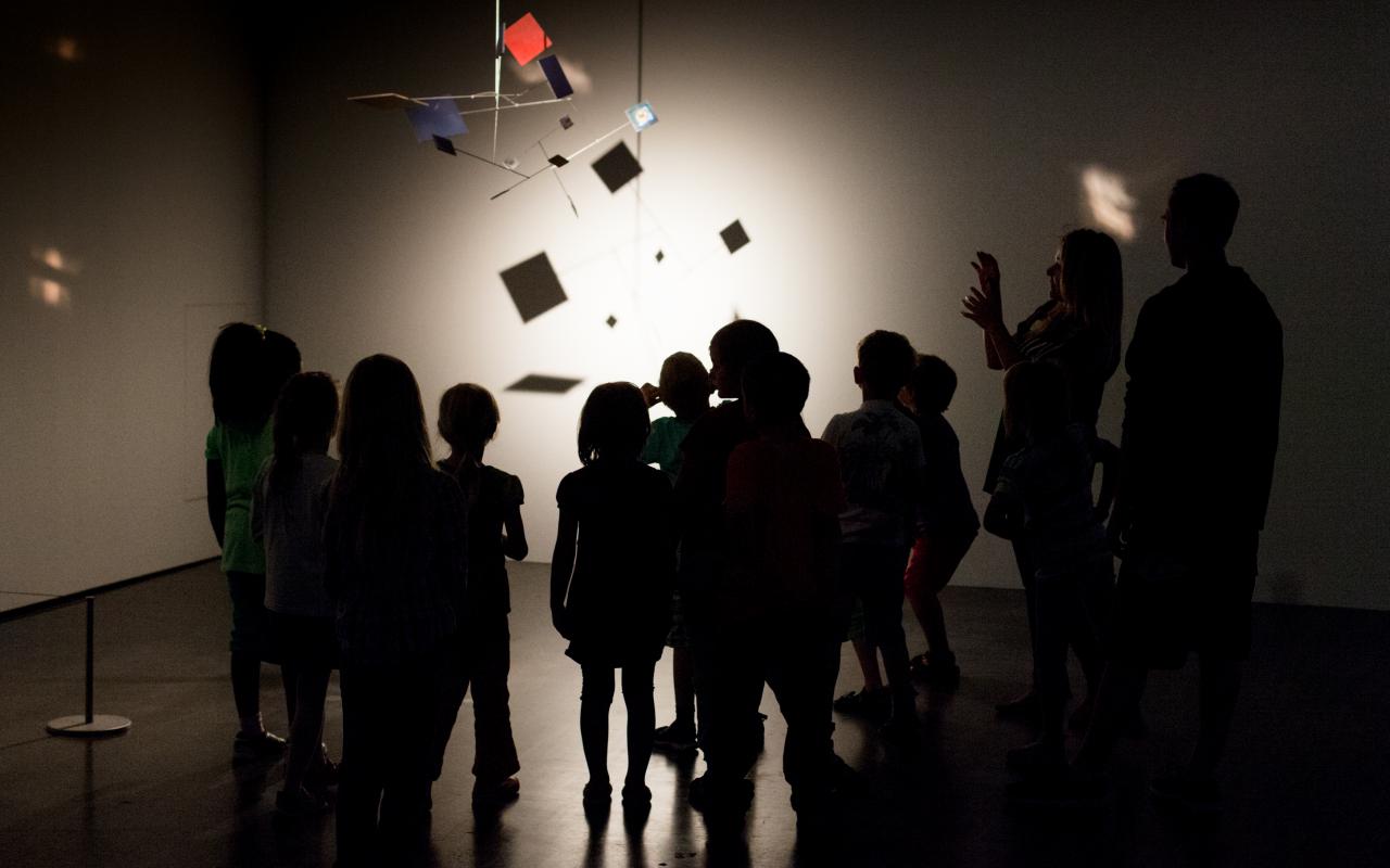 Kids are standing in the dark museum, gazing at an mobile, that is illuminated