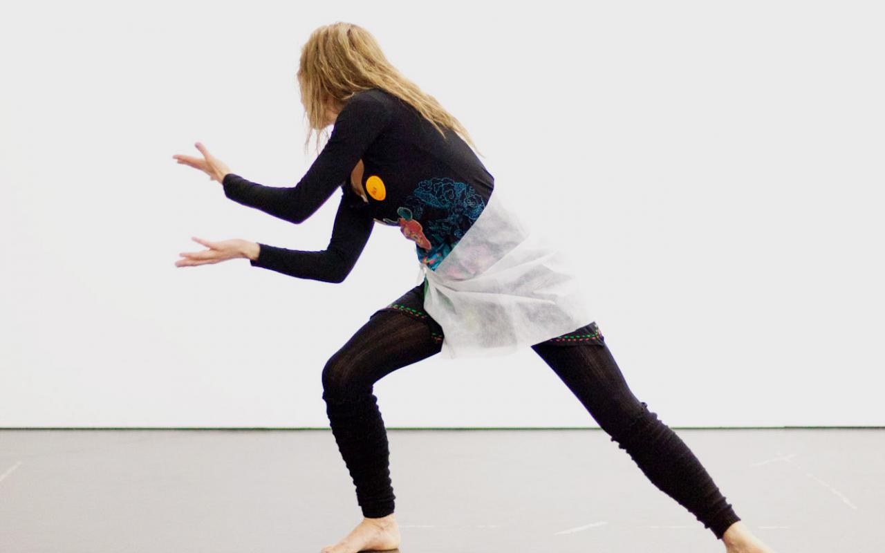 Eine junge Frau in schwarzer anliegender Kleidung ist in tänzerischer Pose abgebildet. Sie hat ihr Gewicht auf das rechte Bein verlagert, das sie angewinkelt hat. Das andere ist in einer Linie mit ihrem Oberkörper ausgestreckt.