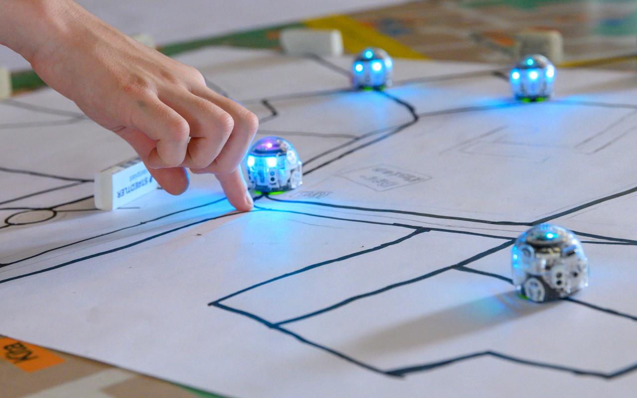 On the photo: Small, blue illuminated robots drive along black lines on a sheet of paper.