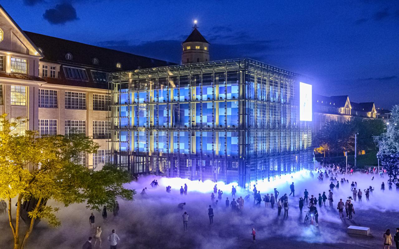 Das Foto ist bei Nacht mit einer Drohne aufgenommen. Es zeigt die Nebelskulptur und viele Silhouetten von BesucherInnen der KAMUNA 2019. Der Kubus und die Fassade des Gebäudes sind hell erleuchtet.  