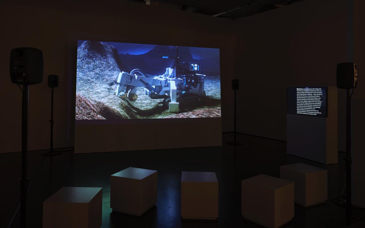 Das Bild zeigt einen Raum mit Sitzkästen, Monitoren und einem großen Bildschirm auf dem der Film »Prospecting Ocean« von Armin Linke abgespielt wird.