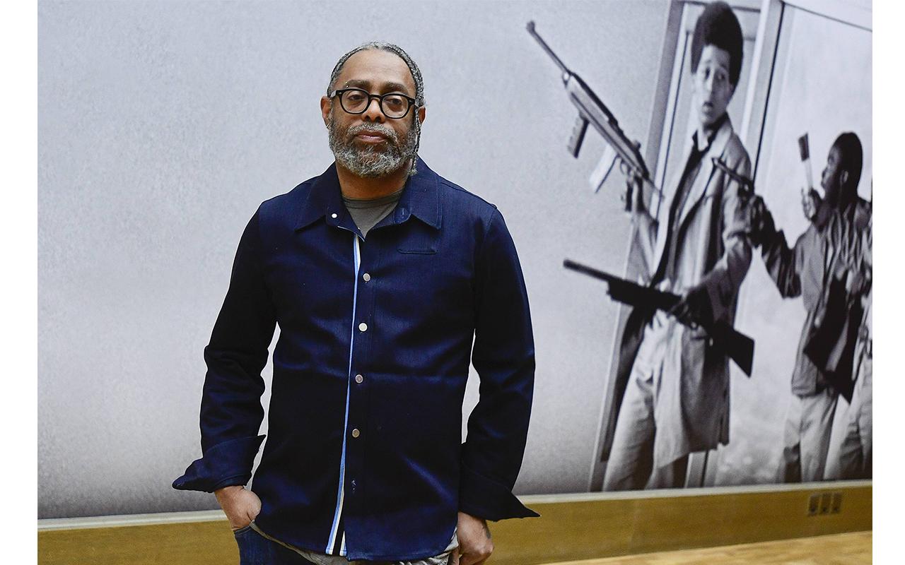 Arthur Jafa steht vor einer Wand, auf der eine Fotografie mit zwei jungen Afroamerikanern mit Waffen abgebildet ist.