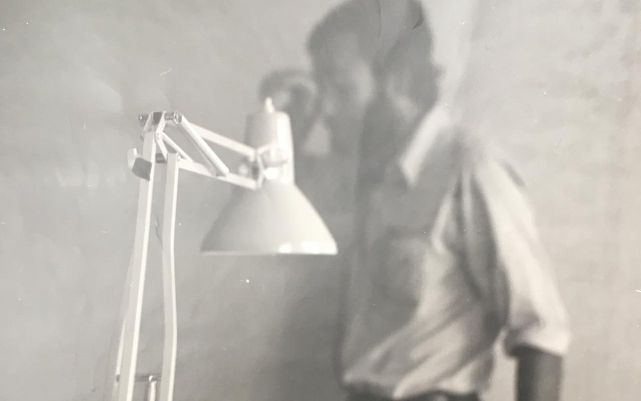 Eine Schwarz Weiß Fotographie zeigt im Vordergrund scharf eine Schreibtischlampe. Im Hintergrund sind man unscharf einen Mann mit Bart und einem weißen Hemd. Es ist Hansjörg Mayer.
