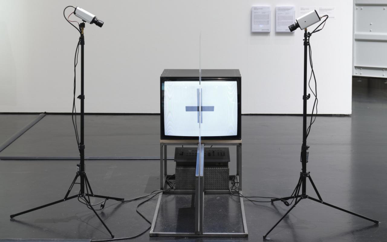 The photo shows a tube screen framed by two cameras. The cameras are mounted on two stands. In the middle of the screen a plexiglass plate is placed on a metal frame. A cross shines from the tube screen.  