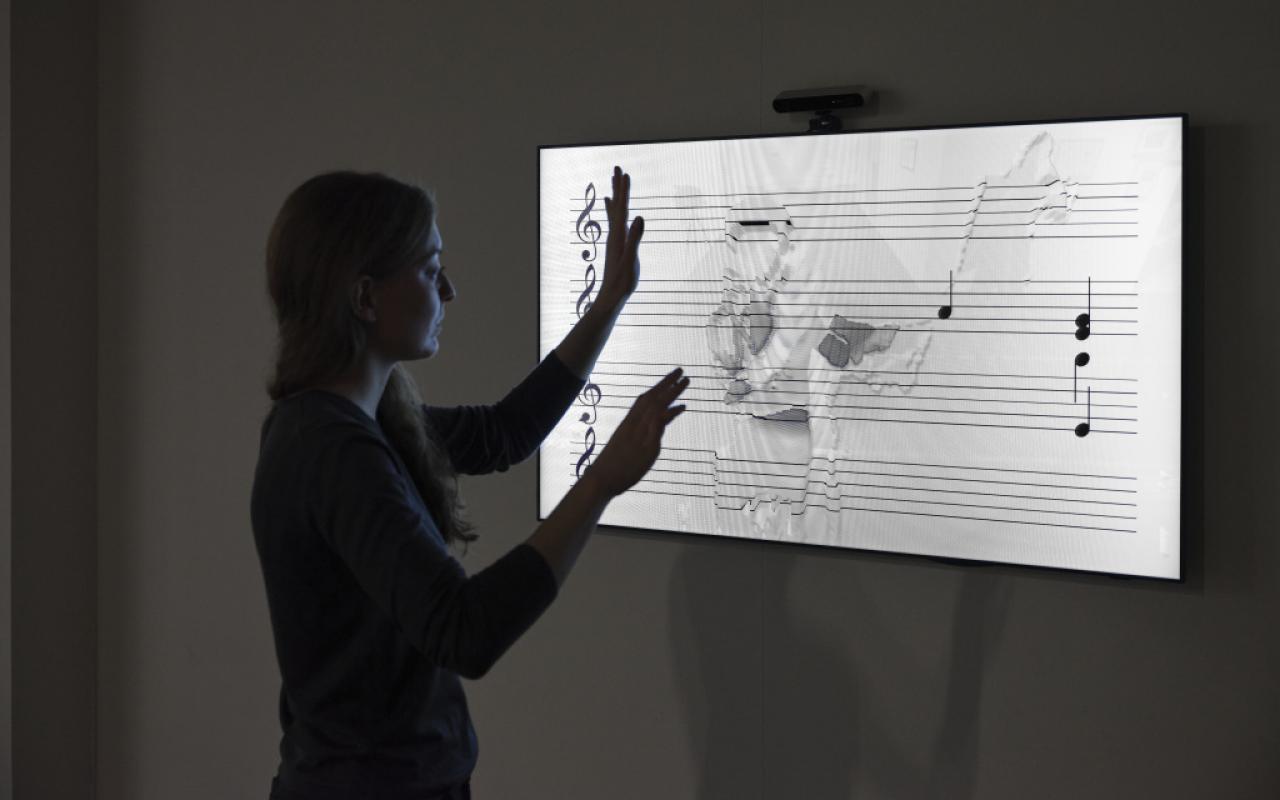 Das Foto zeigt eine Besucherin die mit ihren Händen über eine Noten Patitur fährt. Diese Notenpartitur wird von einem Bildschirm ausgestrahlt und simuliert.