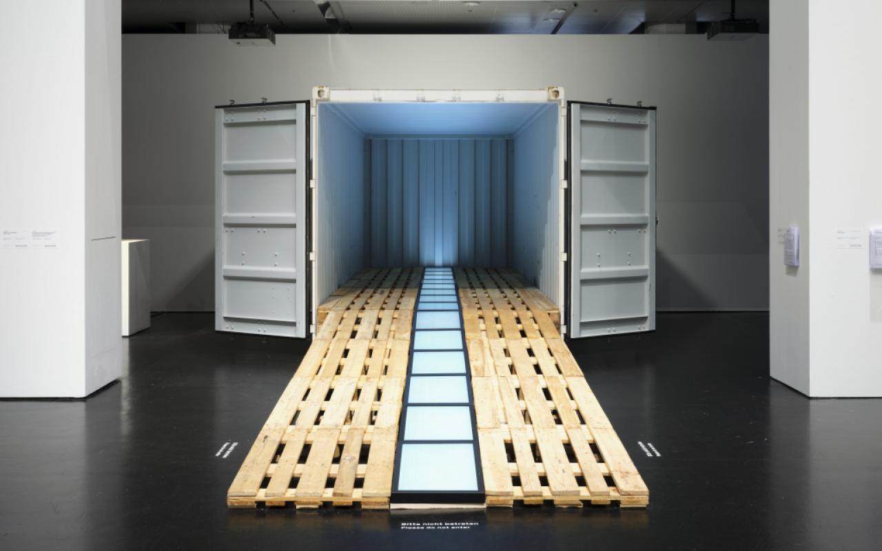 From a container, European panels run out like a ramp. A row of screens runs from the ramp into the container. The container is illuminated in blue.