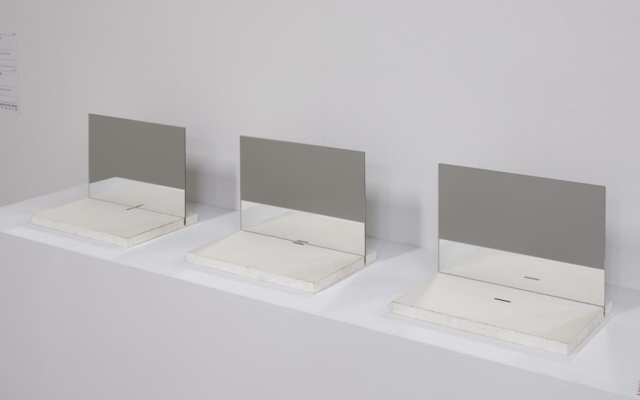 Das Foto zeigt eine dreiteilige Arbeit. Es liegen drei Papiere mit einer jeweils unterschiedlich angeordneten Linie auf Novopanplatten aufgelegt. Vertikal stehen Spiegel auf den Platten und spiegeln die dünne Linie auf dem Papier. 