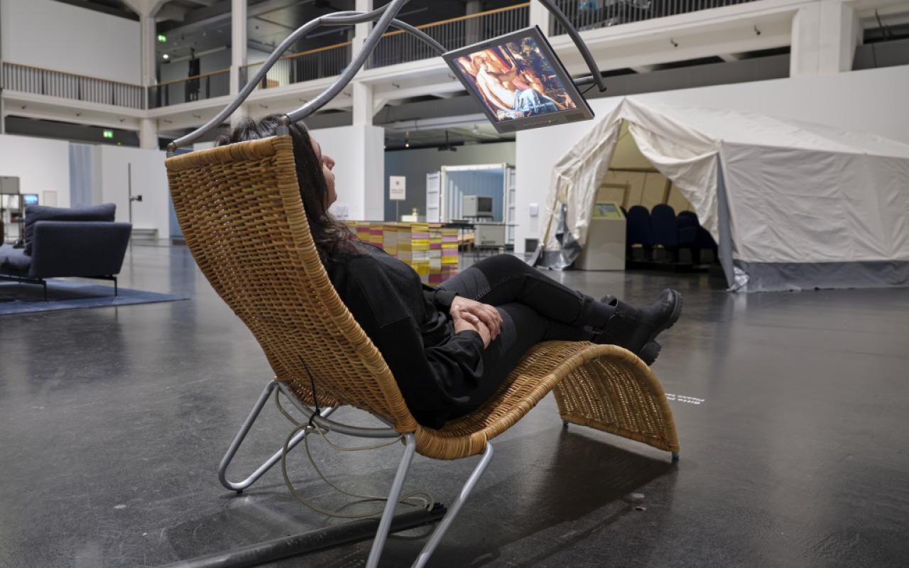 The photo shows a woman lying on a rattan couch. Above the couch is a metal frame which allows a screen to float in the air. On this runs the work Pluriversum by Peter Weibel.