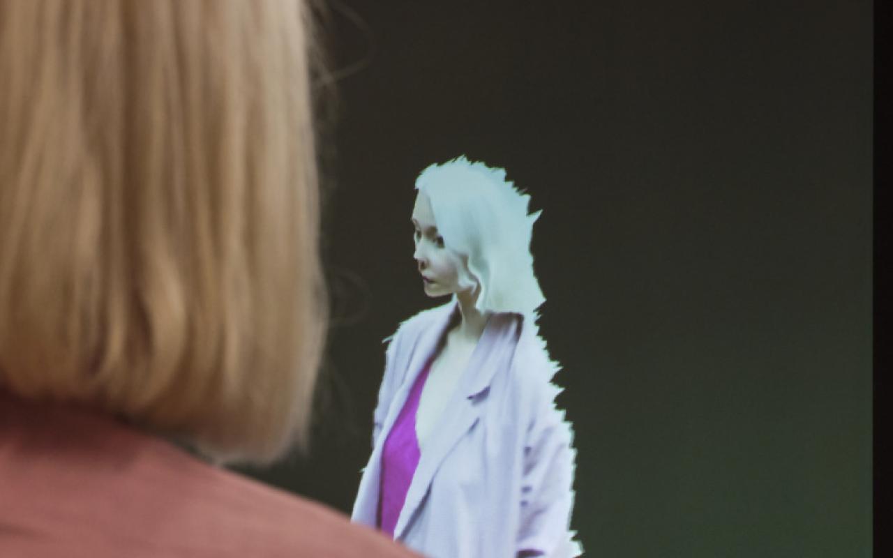 Im Vordergrund des Fotos sieht man eine hellblonde Frau mit kurzen Haaren und einer rosafarbenen Jacke. Vor ihr ist sie als Avatar auf einem Bildschirm reproduziert. Die Besucherin steht ihrem digitalen Code gegenüber.