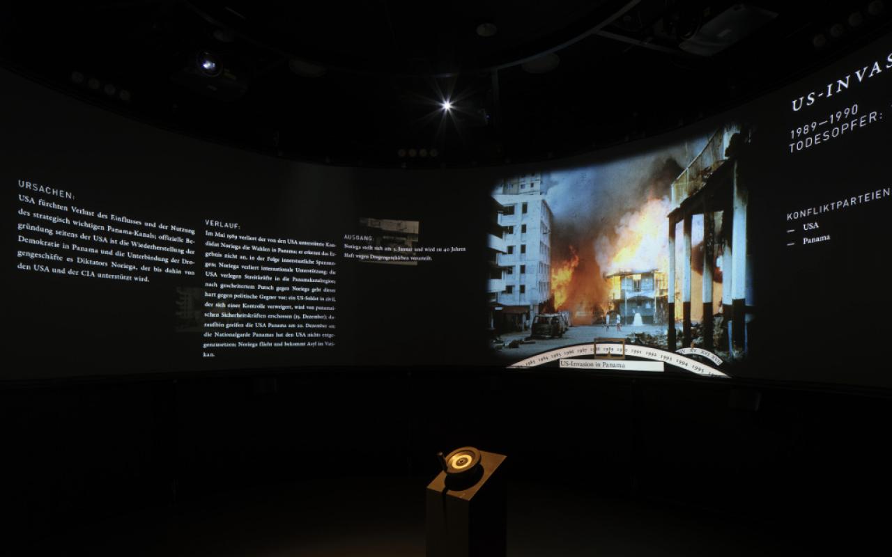 Das Foto zeigt einen verdunkelten Raum, der auf einer großen Panoramaleinwand Kriegsszenarien präsentiert und mit Text ihre Ursachen aufzeigt.