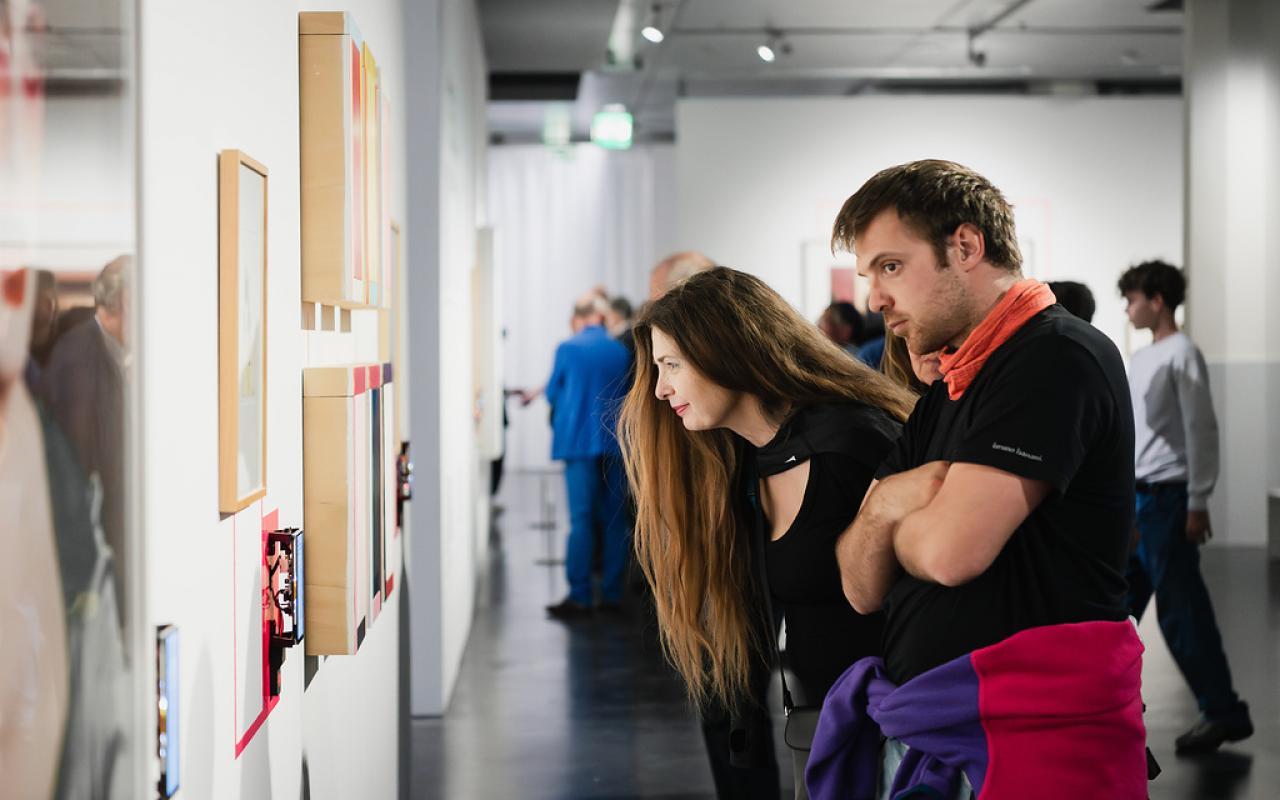 Eine Frau und ein Mann betrachten Bilder an einer Wand. Im Hintergrund sieht man mehrere Menschen durch die Ausstellung laufen.