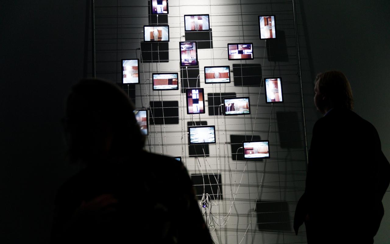 On a metal fence in front of a grey wall hang some tablets which are used as displays. The arrangement is striking, reminiscent of the classic Petersburg hanging. The wiring of the tablets is also notable. A person stands in front of the installation.
