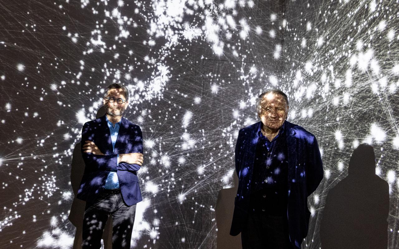 Zwei Männer stehen im Ausstellungsraum vor einer Projektion, die den Kosmos als Netzwerk zeigt. Sie sind von der Projektion überdeckt. Der linke Mann ist der Wissenschaftler Albert-Lázsló Barabási. Der rechte Mann ist Peter Weibel. Beide lächeln.