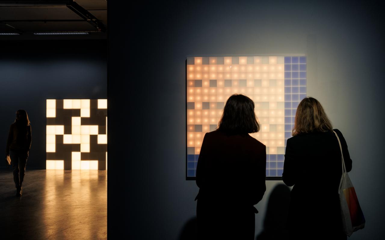 Zu sehen sind zwei Personen vor einem leuchtenden Walter Giers Werk. Im Hintergrund ist ein weiteres würfelförmiges, leuchtendes Objekt zu sehen.