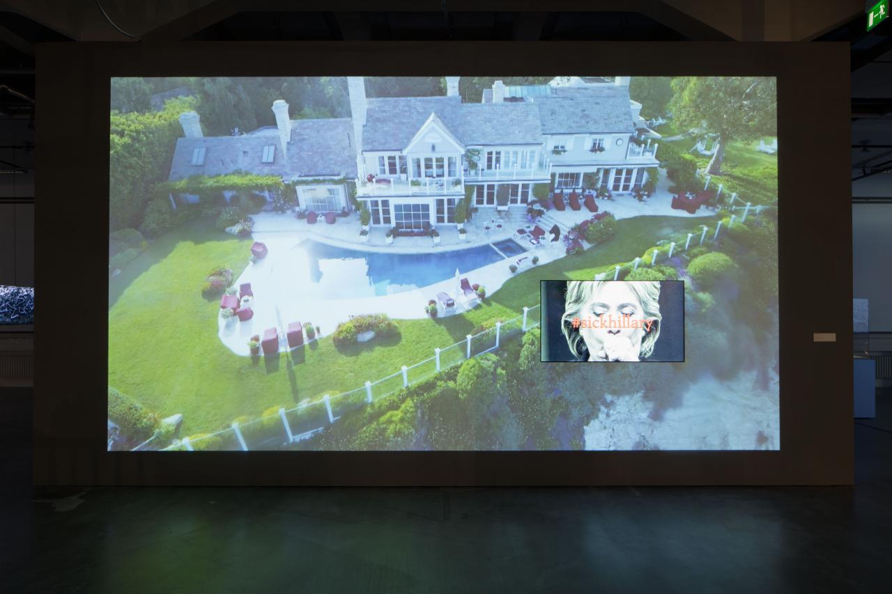 Auf einer Videoleinwand ist eine Villa mit Pool und ein Bild von Hillary Clinton unter dem »sickhillary« steht, zu sehen.
