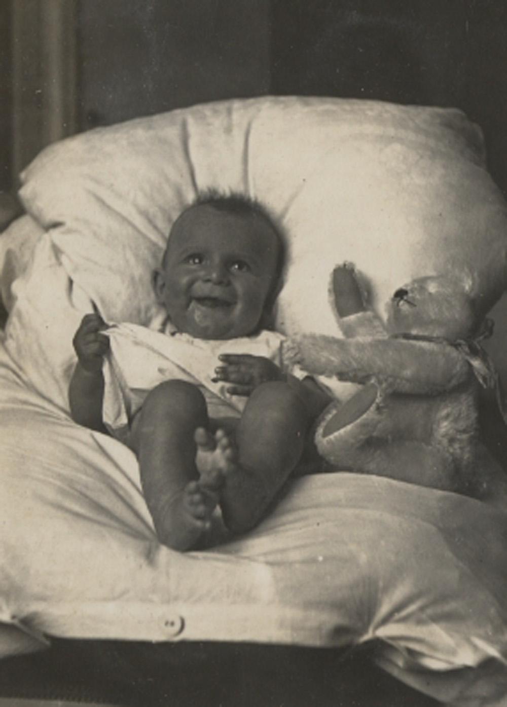 Ein Baby mit Teddybar, auf einem weissen Kissen liegend. 