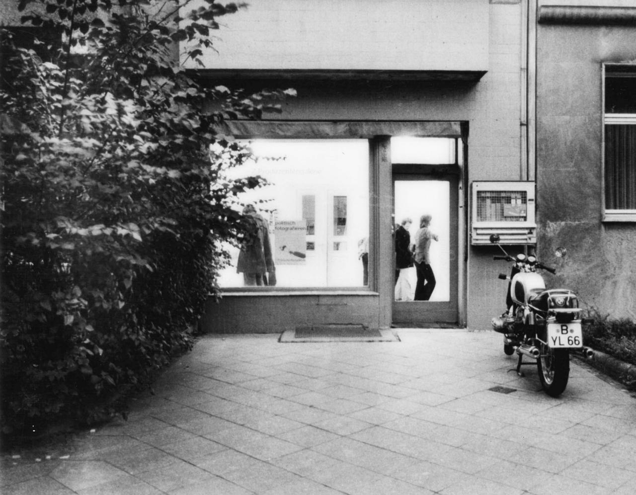 Black and white photo shows the street view of a gallery space. In front of it is a motorcycle.