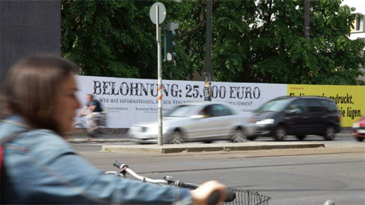 Frau auf einem Fahrrad fährt an einem Plakat vorbei auf dem steht "Belohnung 25000 Euro"