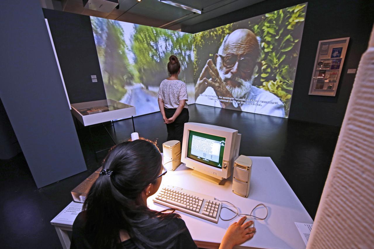 Ausstellungsansicht: ein Mädchen am Computer, im Hintergrund eine wandfüllende Videoprojektion mit Vilém Flusser