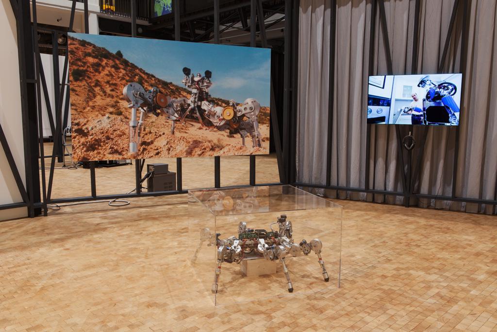 Ein Roboter steht im Vordergrund auf dem Boden, er wird von einer Glashaube abgedeckt. Ein großformatiger Fotodruck und ein Monitor mit Kopfhörern hägen an der Wand.