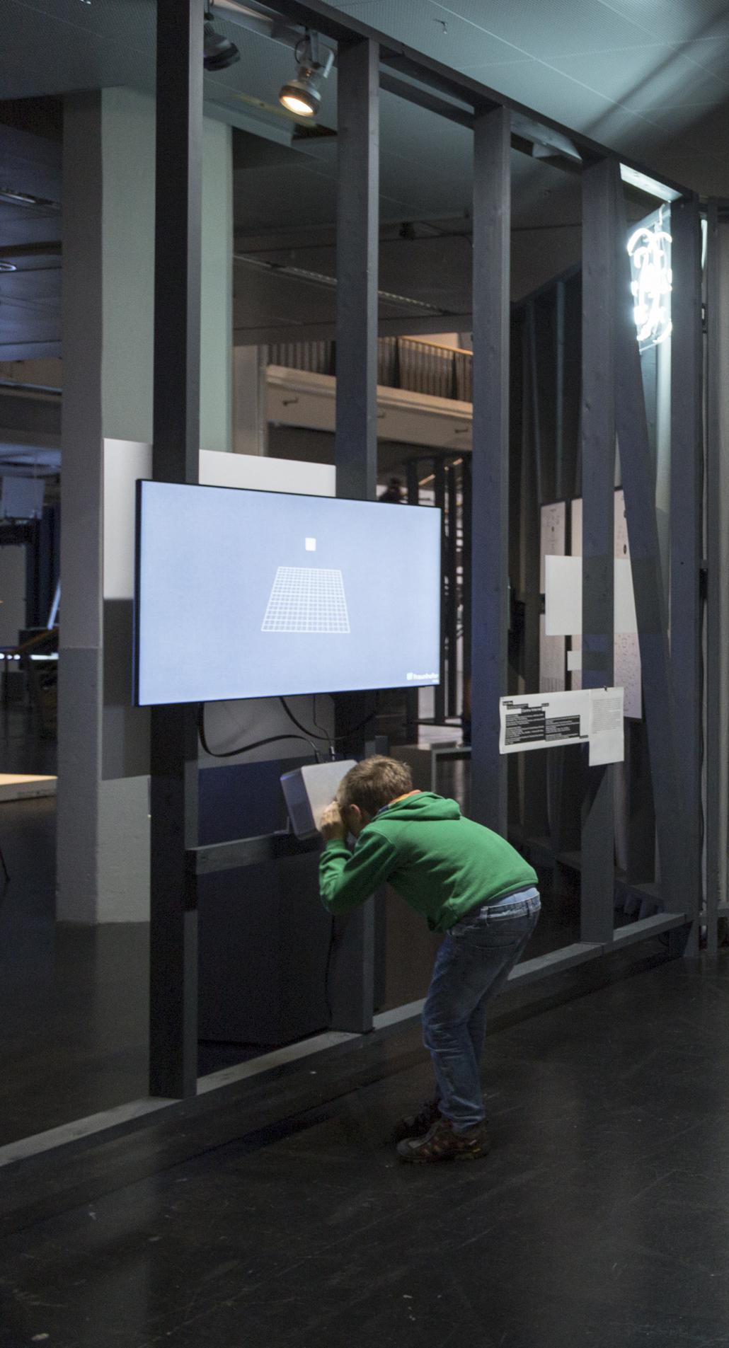 A boy looks through a box. At the wall you can see a screen with a grid box