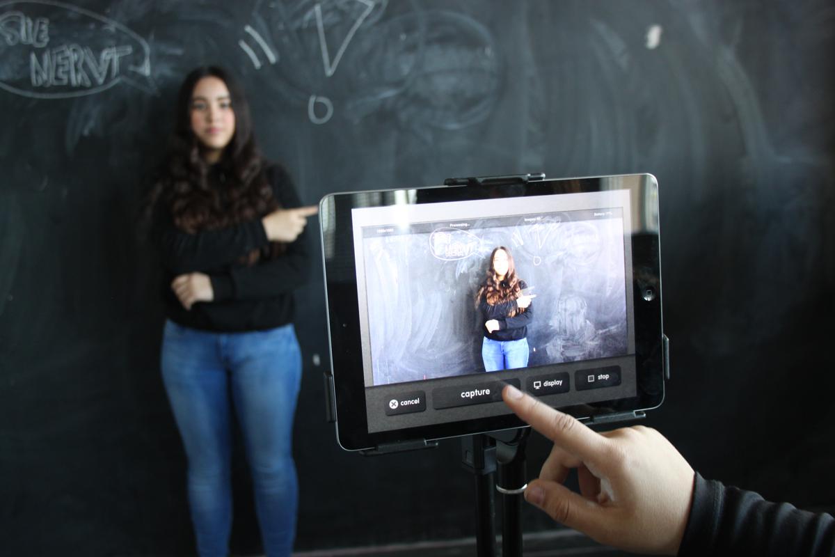 A girl standing in front of a board is being photographed with a tablet. 