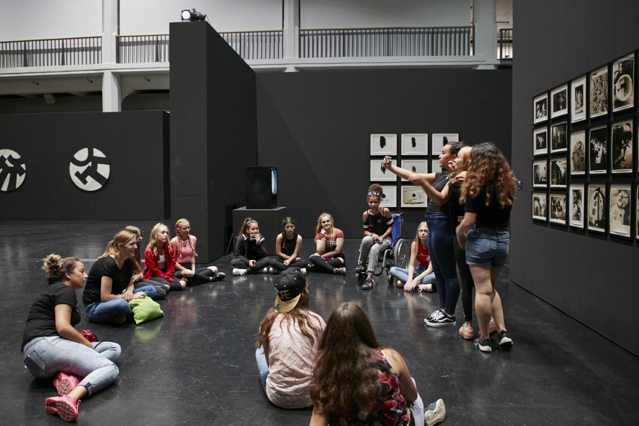 Eine Gruppe von Mädchen sitzt im Kreis in einer Ausstellung. Drei von ihnen machen ein Selfie vor einem Kunstwerk.