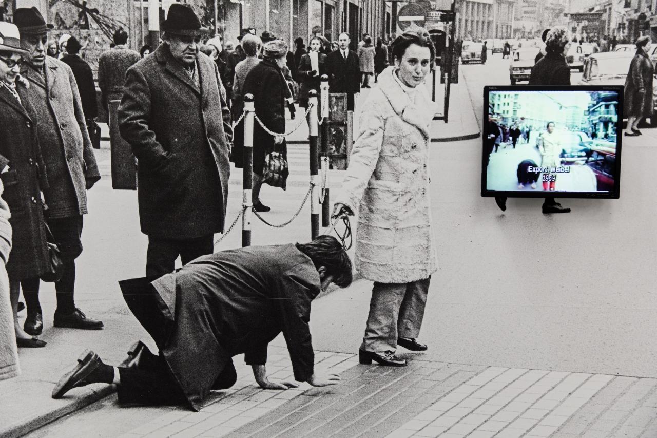 Ein schwarz-weiß Foto: Darauf ist Valie Export zu sehen, die Peter Weibel an der Leine durch eine Fußgängerzone führt.