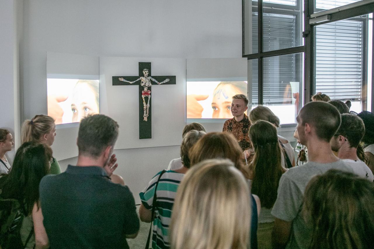 An der Wand hängt ein Kreuz mit einem Gerippe, als Triptychon werden daneben zwei Videos gezeigt. Vor dem Kunstwerk steht eine Menschenmenge.