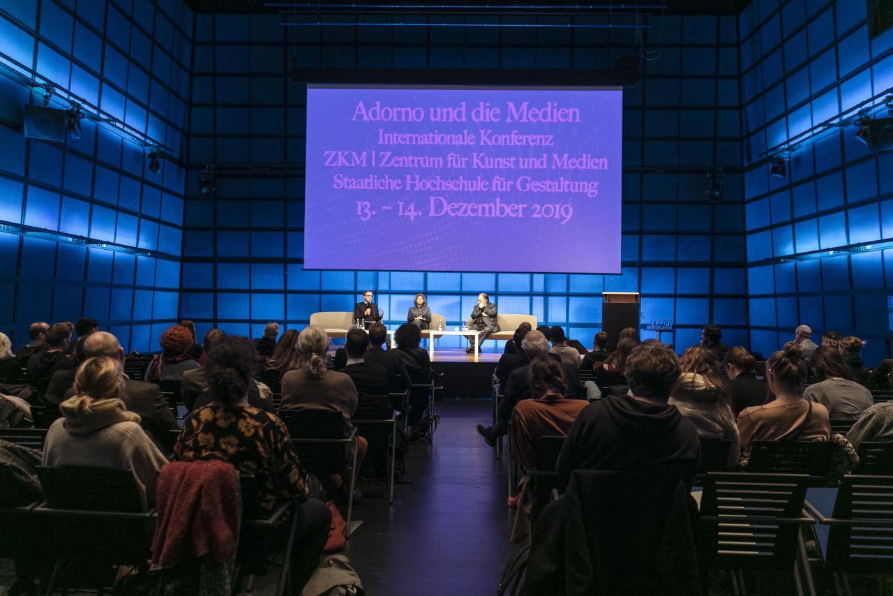 Im abgedunkelten Medientheater erleuchtet das Plakat der Adorno und die Medien Konferenz. Auf dem Podium sitzen von links nach rechts Johan Hartel, Loudmila Voropai und Peter Weibel