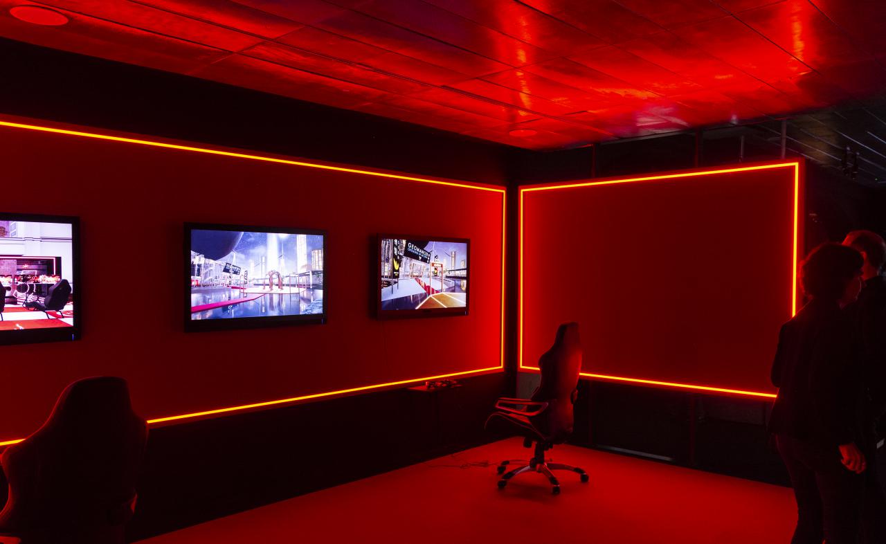 The photo shows a red illuminated room with elegant large office chairs. Three monitors are mounted on the wall, with the chairs in front of them.