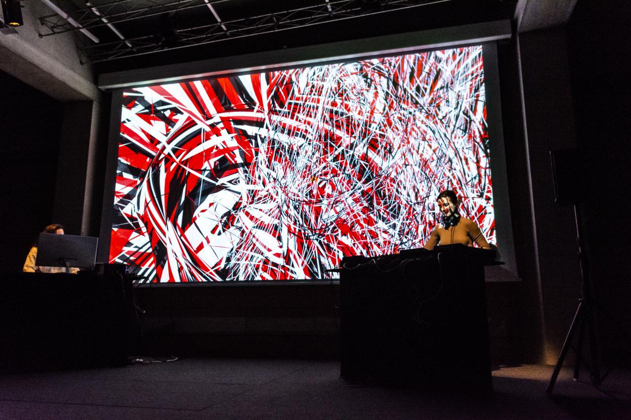 A visual-sound performance will be shown. Two women are standing at the bottom corners of the large screen at their sound controllers while in the background a colour strip of red, black and white merge into one another.
