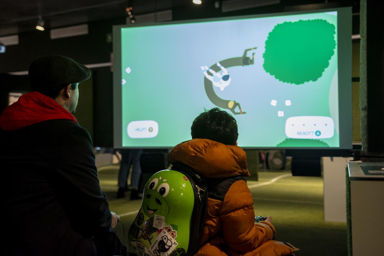 A man and a small boy are turned with their backs to the viewer. The boy is wearing a pear-shaped backpack, which embodies a computer game character. At the back of the picture there is a screen with a game, which is played. 