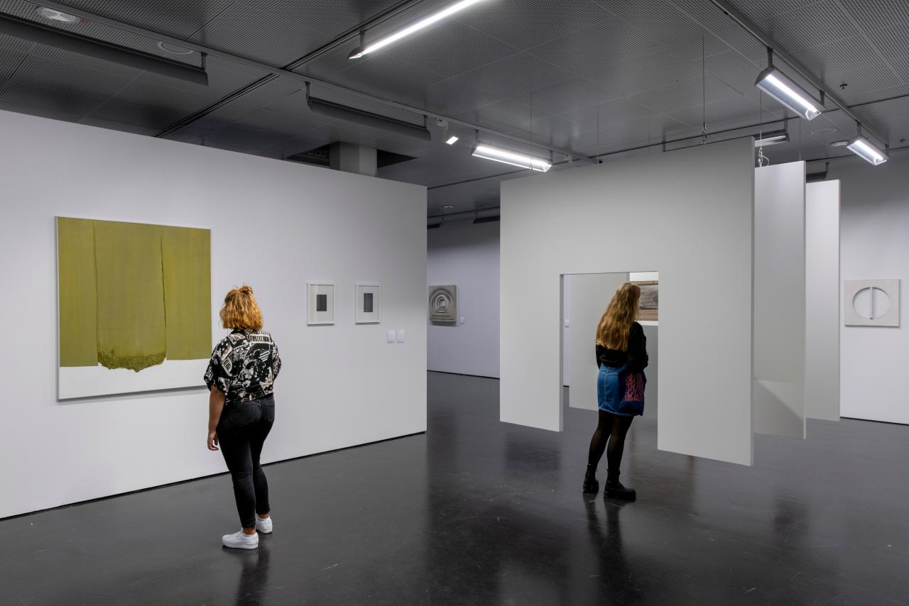 Two women in the exhibition room. On the left a woman is looking at a monochrome painting on the wall. In the middle, a woman is standing in front of a hanging construction consisting of three successive surfaces, with a passageway in between.