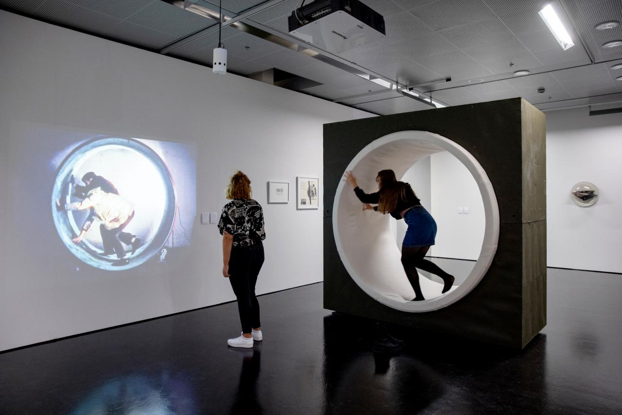 Zwei junge Frauen sind im Ausstellungsraum. Eine betrachtet eine Projektion eines künstlerischen Films an einer Wand und die andere befindet sich in einer Lauftolle.