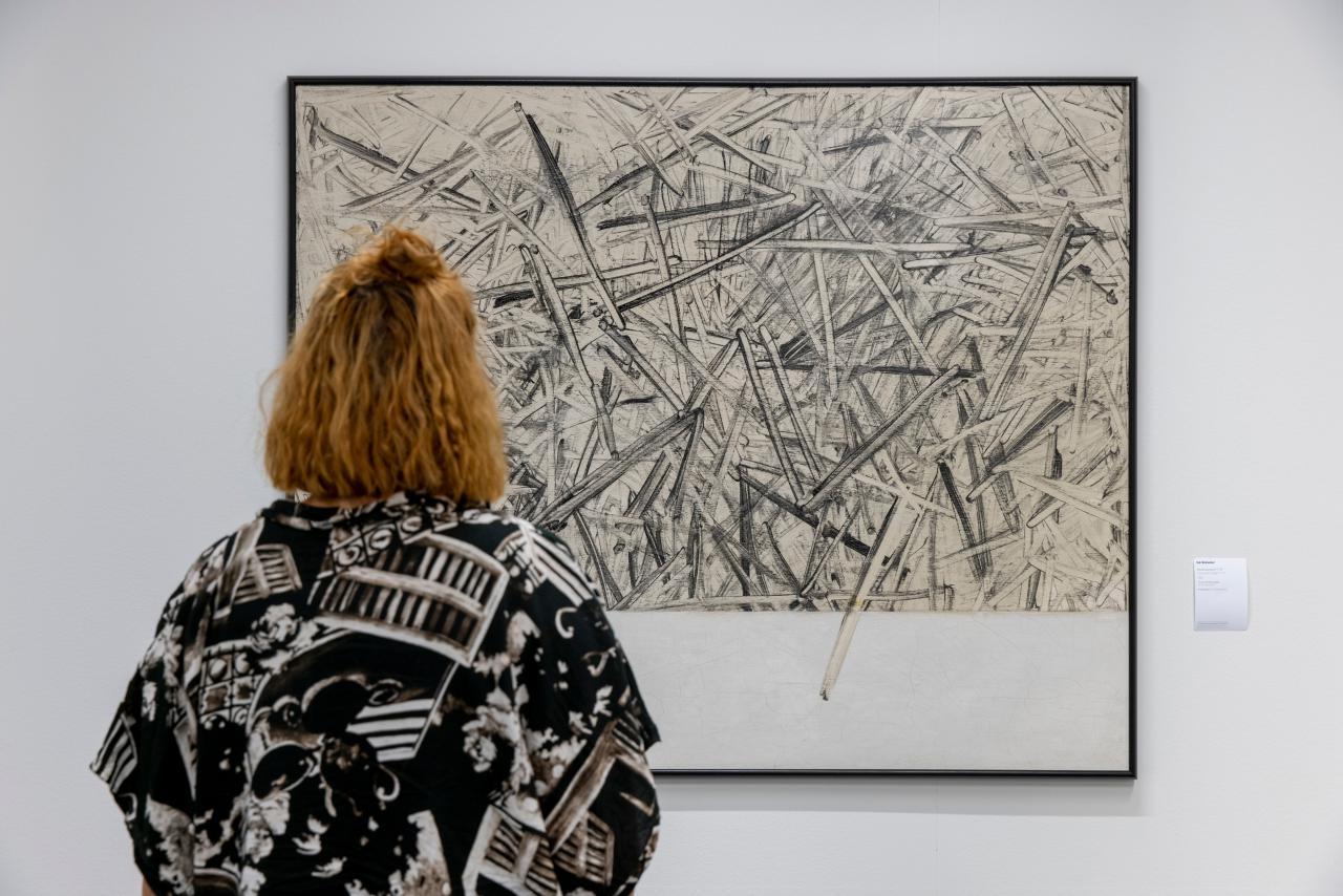 A woman is looking at a picture hung on the wall with sharp thick strokes chaotically painted on top of each other.