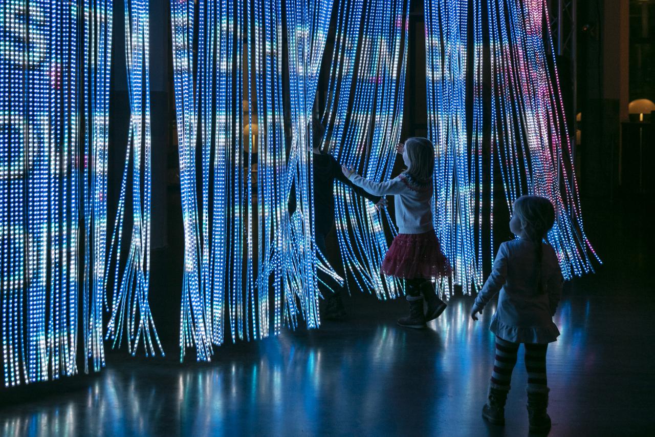 Kinder treten durch einen Vorhang aus Leuchtstäben auf die viele bunte Farben und Schriftzüge projektiert werden.
