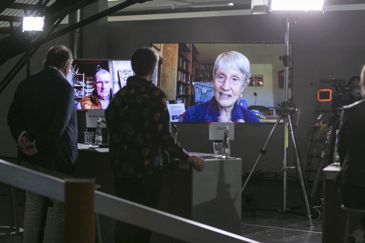 Donna Haraway ist auf einem großen Bildschirm zu sehen. Auf einem weiteren großen Bildschirm ist Bruno Latour zu sehen.
