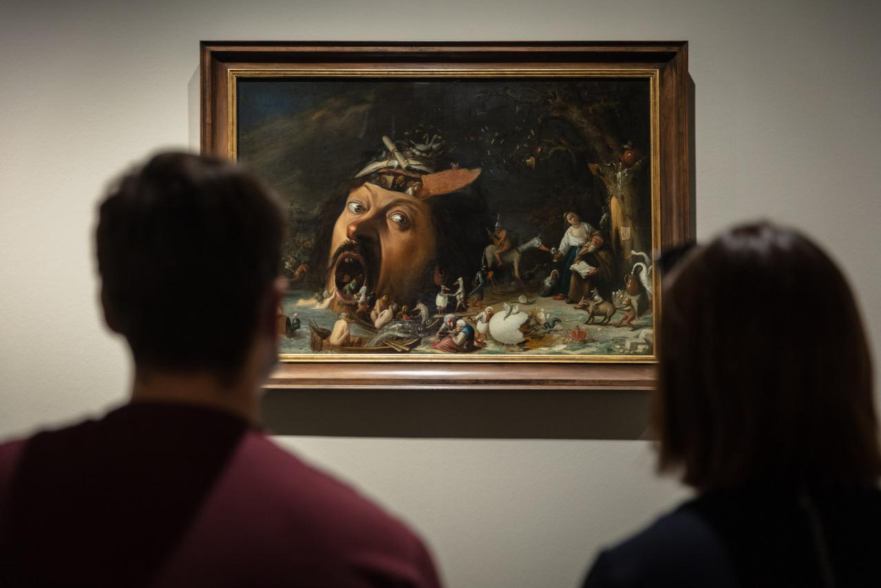 Two people are sitting in front of a picture that shows a huge head with people coming out of its head and mouth. Everything seems very gloomy.