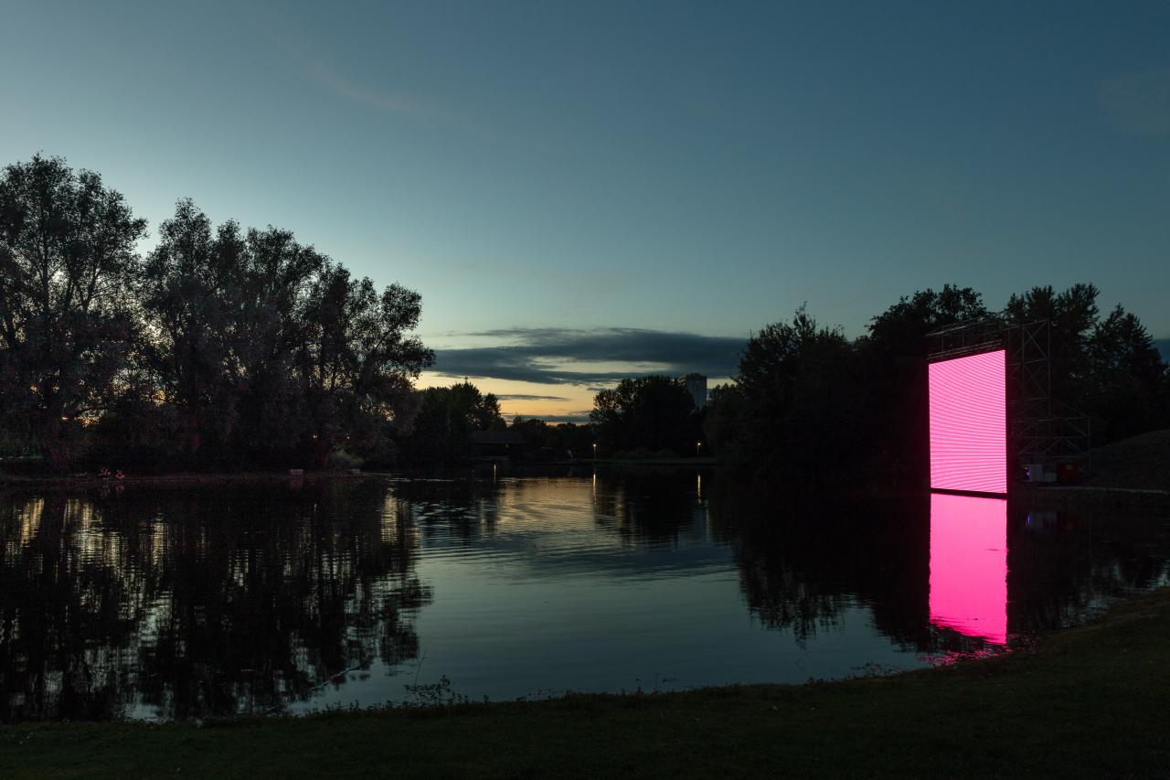 A large square area can be seen, which seems to stand on the surface of a lake in the dark of the night. On the surface the flickering of a screen can be seen. 