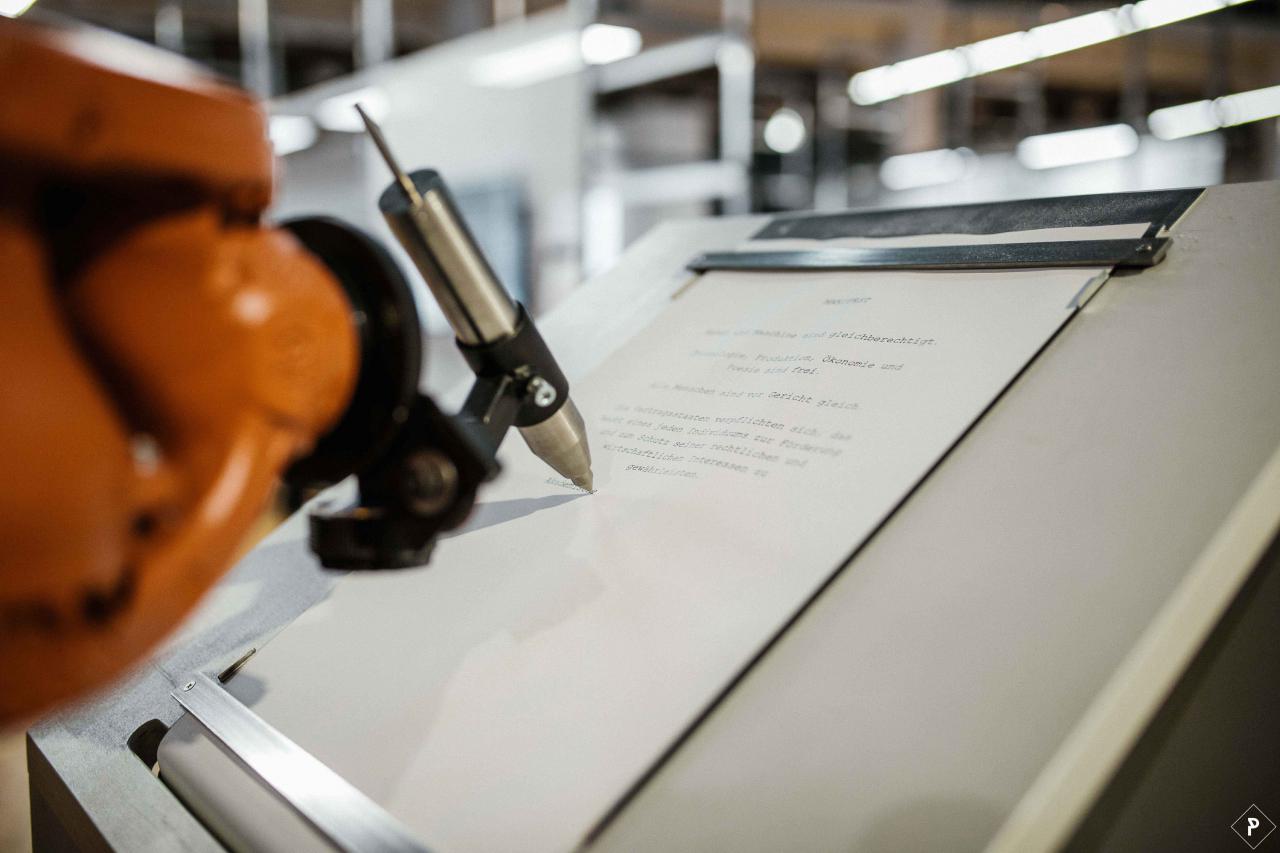An orange robot arm writes with a silver pen on a sheet of paper