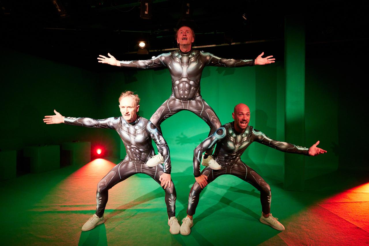 Three men in futuristic armor make a human pyramid in a green screen studio
