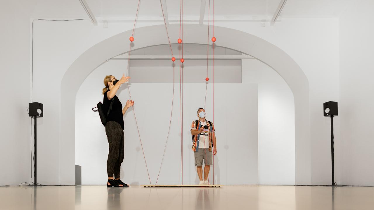 Ein Mann und eine Frau stehen vor einem Kunstwerk, das aus Seilen besteht, welche zwischen der Decke und dem Boden gespannt sind. Die Besucher ziehen die Seile auseinander.