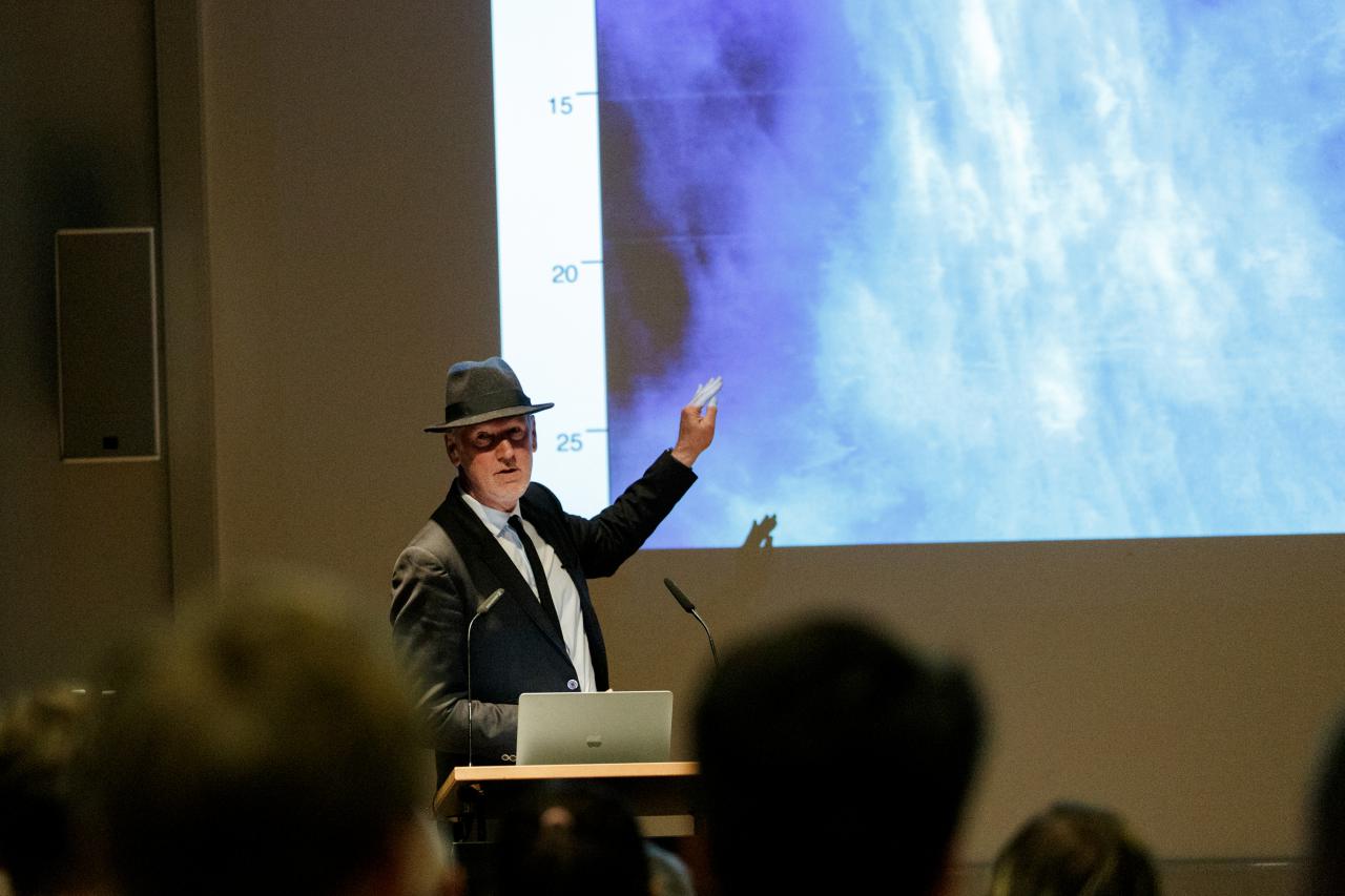Zu sehen ist Thomas Paul, Medienkünstler und Professor in Anzug mit Hut, wie er hinter einem Rednerpult mit Laptop steht und auf die Beamer-Projektion zeigt.