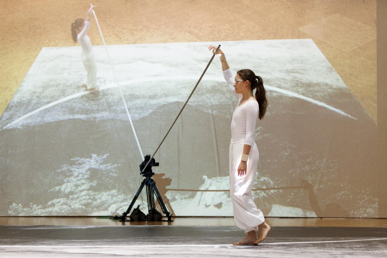 Performance von Ulrike Rosenbachs »Die einsame Spaziergängerin«, dir Künstlerin, ganz in weiß gekleidet hält eine Stange fest und läuft im Halbkreis einen Weg ab, während sie gefilmt wird.