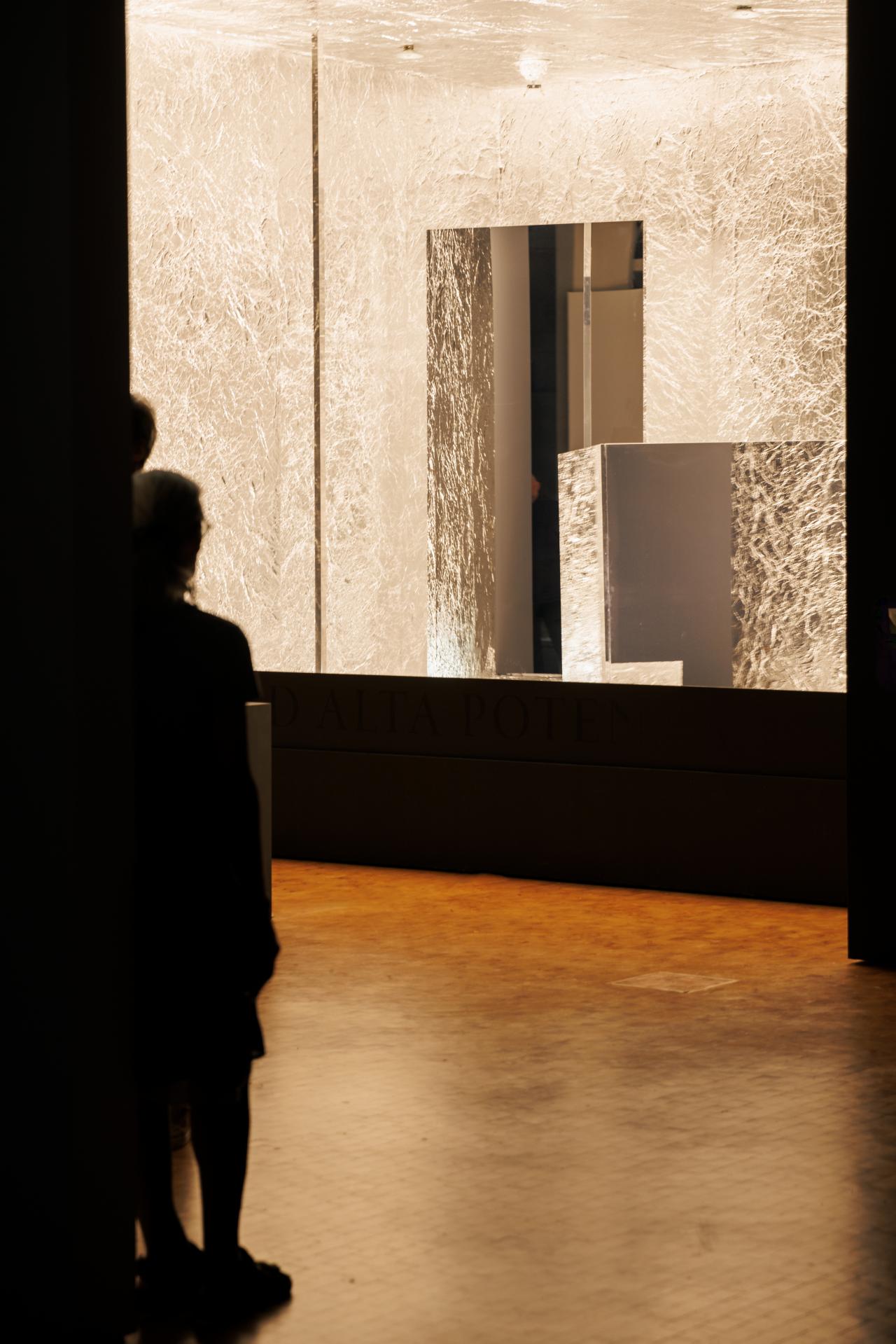 Zu sehen ist ein dunkler Raum, in dem ein helles Kunstwerk steht, das eingelassen in die Wand erscheint und durch Lichter und Spiegel hell ausgeleuchtet wird.
