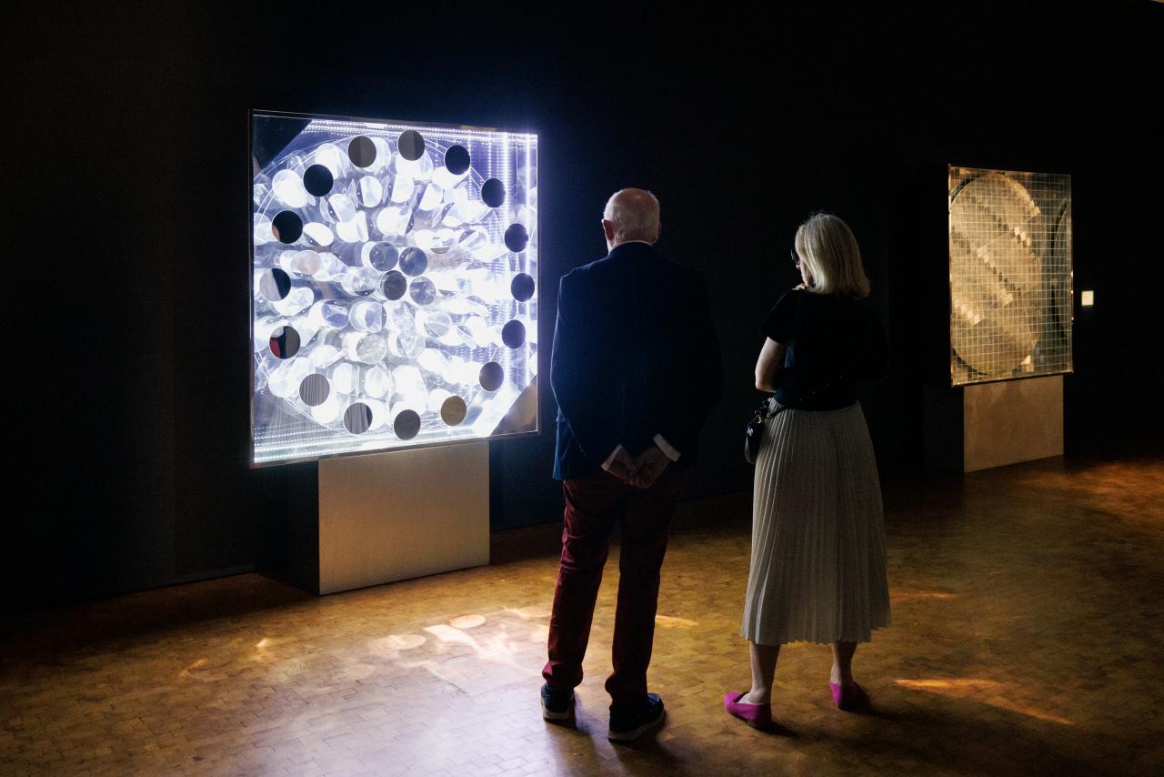 Zu sehen sind zwei Menschen, die vor einem Leuchtkasten stehen, der an der Wand hängt. Durch verschiedene Objekte in den Kasten bricht das Licht und es entstehen optische Variationen.