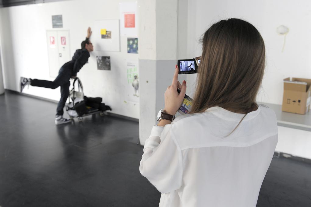A young woman is fotografing a man. He is posing for the picture and is feigning to crash into the wall while pushing a dolly.